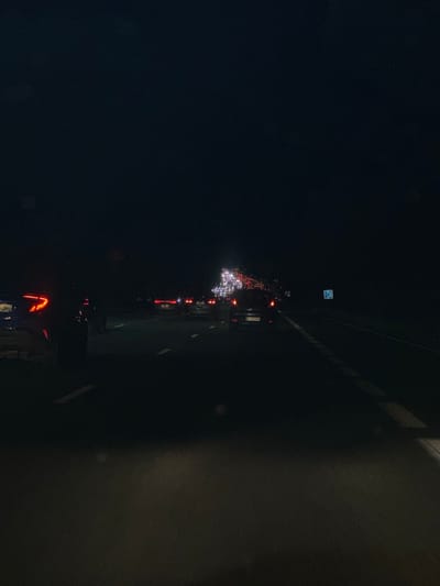 A nighttime, forensic view of a congested multi-lane highway or major road in Saint-Cyr-l'École, France. The perspective is from within a vehicle, looking forward. The scene is dominated by darkness, with illumination primarily from vehicle lights. Directly ahead, numerous vehicles are either stationary or moving slowly, their red taillights forming a dense cluster extending into the distant horizon. Beyond the red taillights, some white headlights are discernible. The asphalt road surface features visible white dashed and solid lane markings. A vehicle with distinct angular red taillights is partially visible in the immediate left foreground. Another vehicle further ahead in the middle lane displays a French license plate, indicating a regional code consistent with Île-de-France (e.g., 91 or 95). On the right shoulder of the road, a blue rectangular sign with a white pictogram is present. The sky above is dark, indicating night, with no discernable environmental features beyond the road and vehicle lights. No individuals are visible, and the primary action is implied traffic congestion. The specific location is on a major road, likely the A12 autoroute or N12 national road, traversing or immediately adjacent to Saint-Cyr-l'École.
