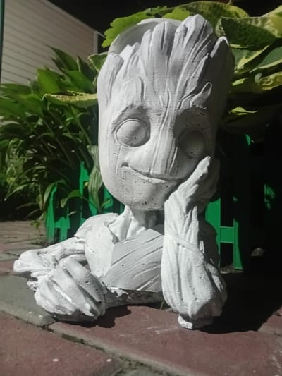 A grey, concrete statue resembling the character Groot from Guardians of the Galaxy is positioned outdoors on a brick-like pathway. The statue depicts Groot's upper body and head, with its right hand resting against its cheek and its left arm outstretched, resting on the ground. The statue is white and textured, with visible cracks and imperfections. Behind the statue, lush green foliage is visible, with some leaves featuring variegated patterns of green and yellow. A bright green fence runs horizontally in the background, partially obscured by the plants. The lighting suggests it is nighttime or dusk, with a spotlight illuminating the statue from the front, casting a distinct shadow to the right. The overall scene appears to be a garden or outdoor decorative area.