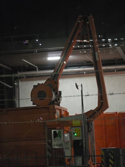 The image depicts a large orange industrial shredder located in Walldorf, Germany. The shredder is a Meinhardt M1085, as indicated by signage on the machine. The machine is in an indoor industrial setting, with pipes, metal grating, and fluorescent lighting visible in the background. The shredder has a prominent rotating drum with jagged teeth, designed for breaking down materials. A control panel with a green illuminated button is attached to the side of the machine. The scene is viewed through a wire fence, suggesting the area may be restricted. The overall impression is of an operational industrial facility.