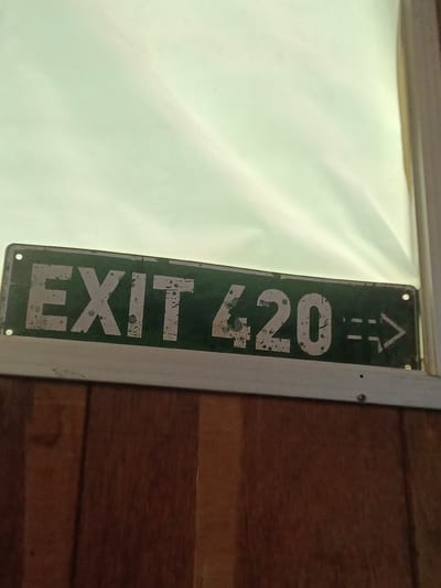 An "EXIT 420" sign is affixed to a brown paneled wall near a window with white curtains. The sign is green with white lettering and a white border. An arrow pointing to the right follows the number "420". The sign is weathered, with some paint chipping and staining. The location is Park Hill, United States. No people are visible.