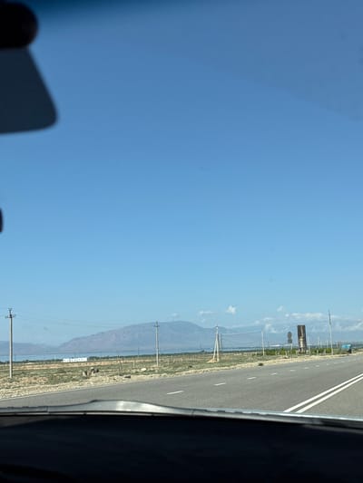 The image captures a view from a vehicle, looking forward on a paved road with white lane markings. The road appears to be in a rural or semi-rural area, possibly on the outskirts of Toru-Aigyr, Kyrgyzstan. The landscape is characterized by a clear blue sky, barren ground with sparse vegetation, and a body of water in the distance, likely Issyk-Kul Lake. Beyond the water, a range of mountains is visible, with some cloud formations.
Along the side of the road, there are utility poles with power lines and fences. In the middle ground, a long, low-rise white building is situated near the lake. Further along the road, to the right, a tall, cylindrical structure, possibly a water tower or an industrial remnant, stands prominently. A traffic sign is visible near this structure. A blue truck is parked on the right side of the road, near the horizon. The foreground shows the interior of the vehicle, including the dashboard and a portion of the windshield frame, with a shadow cast from the rearview mirror. The scene is well-lit, suggesting daylight conditions.