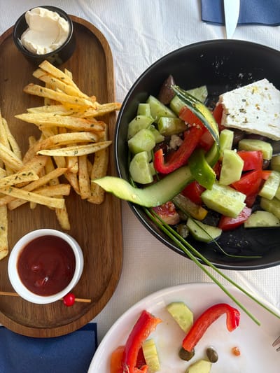 The image shows a close-up view of a table setting with food, likely at a restaurant in Thira Municipal Unit, Greece. Dominating the frame are two dishes: a wooden tray with a generous portion of French fries and a small bowl of mayonnaise, and a black bowl filled with a Greek salad. The salad contains diced tomatoes, cucumbers, olives, red bell pepper, and a large block of feta cheese. A thin slice of cucumber is draped over the salad. To the side, a small white bowl holds ketchup, garnished with a red bead on a toothpick. Partially visible in the foreground is a white plate with leftover pieces of red bell pepper and cucumber, suggesting a meal in progress or recently concluded. The table is covered with a white tablecloth, and a dark blue napkin is visible on the left. The lighting is bright, indicative of daylight, and the overall scene suggests a casual dining experience.