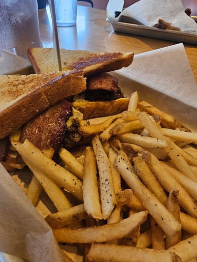 The image displays a basket filled with a sandwich and french fries. The sandwich consists of toasted bread slices, stacked with ingredients that appear to include meat and cheese. The french fries are golden brown and seem to be seasoned with black pepper. The basket is lined with white paper. A skewer pierces through the sandwich. A water bottle is visible on the left side of the image. The setting appears to be a restaurant in Homestead, United States of America.