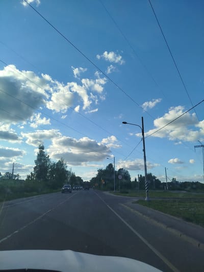 The image shows a street scene in Мамонтово, Russia, during daylight. The road is paved and stretches ahead with several cars visible in the distance. Overhead power lines crisscross the sky, which is blue with scattered cumulus clouds. Roadside vegetation is present on both sides of the road. A speed limit sign indicating "50" is visible. A street light and utility pole are situated on the right side of the road. The photograph is taken from inside a vehicle, as part of the dashboard is visible.