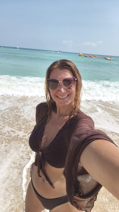 A woman is taking a selfie at a beach. She is wearing sunglasses and a brown bikini. The water is clear and blue, and there are some boats in the distance. The sky is clear and blue. The location is likely not Jiyang District, China.