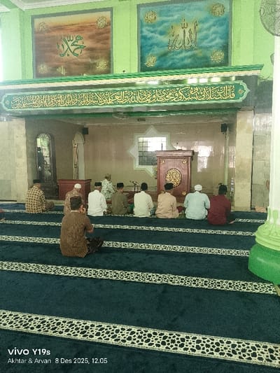 The image captures the interior of a mosque in Jagapura Lor, Indonesia, depicting a scene of religious observance. Several Muslim men are seated on dark blue prayer rugs, arranged in rows, with their backs mostly facing the viewer. They are wearing traditional attire, including kufis (prayer caps) and various shirts, some patterned, some plain. The men are facing towards the front of the hall, where a wooden minbar (pulpit) stands prominently.
Above the minbar and across the front wall, the mosque features light green painted walls adorned with Islamic art. Two large square panels display intricate Arabic calligraphy: the left panel shows the name "Muhammad" and the right panel features the name "Allah," both rendered beautifully against textured backgrounds. Below these panels, a long, ornate band of green and gold Arabic calligraphy stretches across the wall. The ceiling is also light green, and a natural light source is visible through a star-shaped window opening above the minbar.
The activity suggests a congregation attending a religious gathering, possibly a prayer session or listening to a sermon, given their seated posture and focus towards the minbar. The timestamp from the "vivo Y19s" camera indicates "8 Des 2025, 12.05," suggesting the event took place around midday. The atmosphere appears serene and respectful, characteristic of a place of worship. The patterned stripes on the prayer rugs delineate individual prayer spaces.