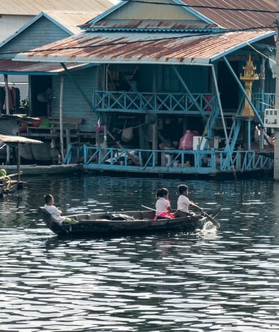 In the city of Soutr Nikom, Cambodia, a boat carries three children on a body of water. The boat is being rowed by a boy in the rear, who is wearing a white shirt and dark pants. Beside him, a girl in a pink shirt and pink skirt sits facing forward. In the front of the boat, a boy in a white shirt and green pants sits facing backward. Behind the boat, a stilted structure, possibly a house or market, is built over the water. The structure has a blue exterior with a rusty tin roof and multiple levels connected by stairs and railings. The water's surface is disturbed by ripples, reflecting the sunlight.