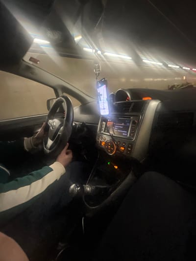 The image depicts the interior of a passenger vehicle from the front passenger's perspective, operating within a brightly illuminated tunnel near Airolo, Switzerland. A driver, wearing a dark garment with white and green arm detailing, has both hands on the black steering wheel, which bears a Toyota emblem. A smartphone, displaying a map application, is mounted centrally on the dashboard and connected by a cable to the car's infotainment system. This system, featuring a silver fascia, shows a digital display with text like "Playback" and "Recommendation," alongside illuminated controls indicating "23.0." A manual gear shift lever is visible in the center console. Partially visible in the lower right foreground is a dark-clad leg belonging to a passenger. The tunnel environment is characterized by continuous linear lighting fixtures along its ceiling and walls, creating bright reflections within the vehicle's interior.