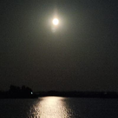 A bright full moon illuminates a body of water at night. The moon, positioned in the upper center of the frame, casts a strong reflection on the water's surface, creating a bright path of light. A dark silhouette of trees or a treeline is visible on the horizon, separating the water from the hazy, dark sky. The overall atmosphere is serene and dimly lit, typical of a moonlit night. This scene is situated in the rural area of Верцялішкаўскі сельскі Савет, Belarus.