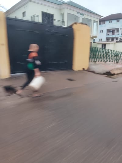 The image depicts a person in motion, walking on a wet paved surface, likely a street in Isheri, Nigeria. The person, wearing dark pants and an orange and green top, is carrying a white cylindrical object in their right hand. The movement blur suggests they are walking at a moderate pace. Behind the individual is a large black metal gate, likely an entrance to a property. To the right of the gate, a yellowish pillar stands, and beyond that, a green metal fence. In the background, light-colored buildings with multiple stories are visible, one of which appears to be a commercial establishment with large glass windows. The overall environment suggests an urban setting with overcast skies, contributing to the muted tones of the image.