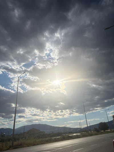 A low-angle, full-shot photograph captures a dramatic sky over a landscape in Prosotsani Municipal Unit, Greece. The sun, obscured by heavy, dark clouds and lighter, wispy formations, shines brightly, creating a radiant halo effect and casting rays across the scene. The sky transitions from dark, stormy gray clouds overhead to lighter, scattered clouds and patches of blue near the horizon.
In the foreground, a paved road with white lane markings occupies the lower right quadrant. To the left of the road, a strip of dry grass and vegetation lines the shoulder, with several tall, modern streetlights interspersed along the roadside. In the middle ground, a residential building with a red-tiled roof and a concrete structure are visible, surrounded by greenery. Beyond the structures, a range of distant, hazy mountains forms the backdrop, silhouetted against the sky. The overall impression is one of a powerful natural spectacle occurring over a rural or semi-rural setting.