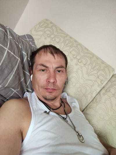A man is lying on a couch. He is wearing a white tank top and two necklaces. One necklace is a thin black cord with a small silver pendant, and the other is a thicker silver chain with a larger oval pendant. He has short brown hair and blue eyes. He is wearing a white wireless earbud in his left ear. He is looking directly at the camera with a neutral expression. The couch has a patterned cushion behind his head and a grey and white plaid blanket next to him. The location appears to be indoors, likely in a private residence. There is no definitive information to confirm the location as Gus-Khrustalny, Russia.