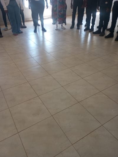 The image displays an indoor scene in Racecourse ward, Kenya, featuring a group of approximately ten individuals standing on a light-colored, square-tiled floor with dark grout lines. Strong ambient light from the background casts reflections on the floor, suggesting a window or bright opening. Only the lower half of each person, from the waist or thigh down, is visible. Attire includes light blue wide-leg jeans with white open-toed footwear, dark trousers paired with various dark closed-toe shoes (sneakers, boots), and a long, multi-colored patterned dress with white sneakers. The individuals are stationary, positioned in a loose semi-circle, with no discernible actions or direct interactions occurring.