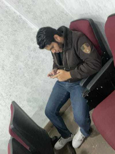 A person with dark hair sits between rows of theater seating, appearing engrossed in their phone. Dressed in a brown hooded sweatshirt and blue jeans, they wear white sneakers. The setting is a theater with red upholstered seats and a speckled floor. The individual is situated under the flipped-up seat of one row while potentially seated in the row behind them.