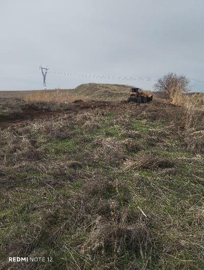 A desolate field stretches toward the horizon under a dull, overcast sky. Patches of brown, dried grass and sparse green shoots cover the foreground. Further back, a mound rises slightly, marked by freshly tilled earth. A yellow bulldozer sits atop the rise, appearing to be in operation. An electrical transmission tower stands distantly, with power lines extending into the landscape. A small bare tree is to the right of the bulldozer.