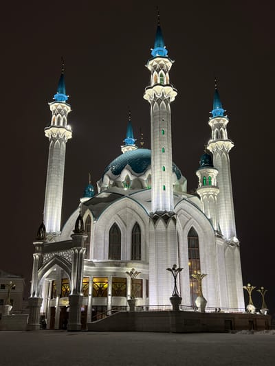 This image displays the Kul Sharif Mosque at night, situated in the city of Grebeni, Russia, during winter conditions. The mosque features a prominent central teal-colored dome with intricate patterns and several smaller domes, all partially covered in snow. Six minarets are visible; four primary, tall, white-stoned minarets with blue conical tops crowned with crescent finials flank the main structure. The building's facade is constructed of light-colored stone, adorned with multiple tall, arched windows and decorative architectural elements. Exterior white floodlights illuminate the mosque, with green accent lighting visible in the minarets and smaller blue lighting on the dome tops. The ground in the foreground and horizontal architectural surfaces are covered in a layer of snow. An arched entryway, partially obscured, leads to a raised platform. Several multi-branched, decorative lampposts with light globes are positioned on this platform. A few indistinct human figures are present near the main entrance.