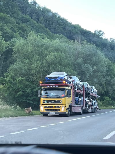 A yellow car carrier truck is traveling on a road in Robești, Romania. The truck is loaded with multiple cars, including a blue sedan on the top deck and several SUVs and sedans on the lower decks. The truck's headlights are on, and the license plate is visible. The truck is positioned on the right side of the road, with the surrounding environment consisting of a lush, green forested hillside to the left and the road stretching ahead. The sky is bright, suggesting it is daytime.
