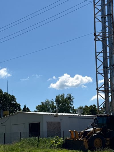 The image depicts an outdoor scene on a bright, sunny day, featuring industrial infrastructure, a building, and a construction vehicle.
At the top of the frame, a large, robust metal truss structure spans horizontally, supporting multiple thick, cylindrical pipes or conduits. Below this, several thin, dark lines, likely power lines, stretch across the sky from the left.
On the right side of the image, a light-colored building, possibly a modular unit or shed, stands with a corrugated metal roof. One window opening on this building is covered with a piece of plywood. Partially visible behind and to the right of the building is a yellow and black construction vehicle, which appears to be a backhoe or front-end loader. The text "605 Super II" is faintly visible on the side of the vehicle.
Green trees and dense vegetation are present around and behind the building and vehicle, suggesting a partially overgrown or natural environment adjacent to the man-made structures. The sky is a clear, bright blue, punctuated by scattered white cumulus clouds, with one prominent, elongated cloud form dominating the upper-middle area. The scene is devoid of human activity, conveying a sense of stillness.