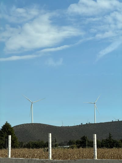 A sunny, daytime landscape shows a blue sky with wispy white clouds. In the distance, wind turbines stand atop a grassy hill, positioned behind a wire fence and concrete posts. Some trees are present on the slope. The image has been rotated, causing the sky to appear on the left and the ground on the right.