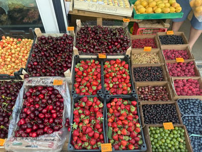 The image shows an outdoor fruit stand in Trubino, Russia, with various berries and cherries for sale. Several cardboard boxes and plastic containers are filled with different types of fruit. From left to right and top to bottom, the fruits visible are: light-colored cherries, dark red cherries, strawberries, red currants, white currants, black currants, raspberries, blueberries, and gooseberries. Some of the fruit containers have price tags with Cyrillic writing and numbers. A person's legs and lower torso, wearing blue shorts, are visible in the background on the right side of the image, suggesting they are the vendor or a customer. The overall scene depicts a typical market stall selling fresh produce.