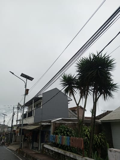 This scene is depicted in Ambakirano, Madagascar, on an overcast day. The image captures a street view with utility infrastructure and buildings. A paved road or pathway is visible on the lower left, bordered by a concrete curb or low wall. A tall metal street light pole stands prominently on the left, fitted with an LED luminaire and a solar panel mounted above it. Numerous black electrical and communication wires crisscross the sky, running diagonally and horizontally across the frame.
Behind the pole, a two-story building features a grey-painted upper facade and a white lower balcony area with a metal roof structure. Below this, a single-story structure with a corrugated metal awning appears to be a storefront or residence. Further down the street, additional buildings with various roof types are partially visible. On the right side of the frame, a cluster of tall, palm-like trees with spiky, dark green foliage rises, partially obscuring structures behind them. Below these trees, a low concrete wall supports a prominent, multi-colored sign with individual letters spelling "662 PASUNDAN RADIO". Green shrubbery grows around the base of the trees and near the sign. In the far left background, a person on a motorbike is faintly visible moving away on the street. The sky is uniformly light grey, indicating heavy cloud cover.