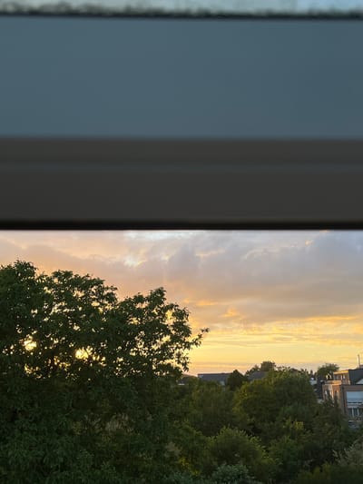 This is a medium shot of a sunset viewed through what appears to be a window or blind. The top of the image is dominated by a dark, horizontal band, likely the window frame or a blind. Below this, the sky is filled with clouds tinged with orange and yellow from the setting sun. In the foreground, a large, leafy green tree is silhouetted against the sky, with sunlight filtering through its branches. In the background, a cluster of residential buildings and more trees are visible. The scene suggests a suburban or residential area in the city of Mondercange, Luxembourg. There are no people or animals visible in the image, and no action is taking place. The overall impression is one of a peaceful evening scene.