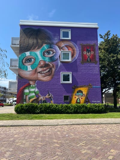 The image depicts a mural painted on the side of a purple apartment building in Leeuwarden, Netherlands. The mural features a large, stylized portrait of a child wearing a superhero mask and cape, with a green and white striped shirt. The child has a wide, toothy grin and large, expressive eyes. To the right of the child's portrait, there are two smaller framed artworks on the wall. The upper frame contains a cartoonish depiction of a monkey dressed as Batman, and the lower frame shows a smaller version of the child character, also with a mask. The building has several windows and balconies. The mural is located on Cornelis Trooststraat, as indicated by a street sign visible in the image. In front of the building, there is a manicured green hedge, a patch of grass, and a sidewalk. The foreground shows a brick-paved street with a herringbone pattern. The sky is clear and blue, and a large tree is visible to the right of the building. Cars and other buildings can be seen in the background.