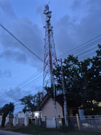 The image shows a telecommunications tower in Pantegurah, Indonesia. The tower is red and white and has several disc-shaped antennas at the top. Power lines run from the tower to the nearby buildings. The sky is cloudy and it appears to be dusk or dawn. There are buildings and trees in the background. The road is at the bottom of the image, indicating the perspective is from ground level.