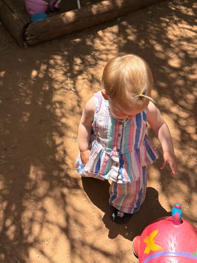 A high-angle shot captures a young child in Misgav Regional Council, Israel, positioned on a dirt surface dappled with shadows. The child has light-colored hair pulled into a small ponytail with an orange hair tie. They are wearing a striped shirt dress. To the right of the child is a red toy. A wooden post is visible on the left edge of the frame. The ground is dry and textured with dirt.