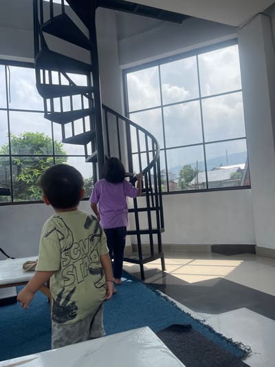 Two children stand inside a building with a large window. One child wears a green shirt with a dinosaur design and gray shorts. The other child, wearing a purple shirt and blue pants, stands on a black metal spiral staircase, apparently taking pictures of the surrounding scenery with a smartphone. Outside, a view of what is likely Sumberdalem, Indonesia, can be seen, featuring buildings and trees under a partly cloudy sky. A blue rug lies on the floor near the children.