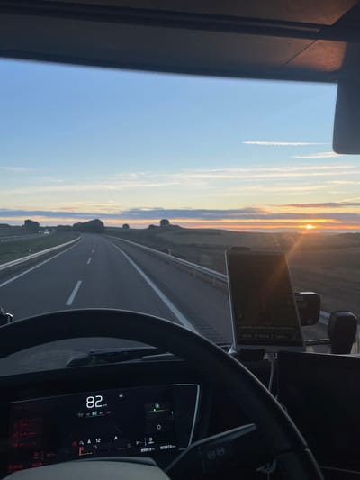 A car dashboard view reveals a highway under a sunset sky with contrails. The speedometer reads 82 km/h, with a mileage of 99999.9 km and a total distance traveled of 303970 km. A GPS navigation system shows a route. The vehicle is traveling on a paved road, flanked by open landscape under a sky painted with hues of orange, yellow, and blue. The steering wheel frames the scene, suggesting the driver's perspective.