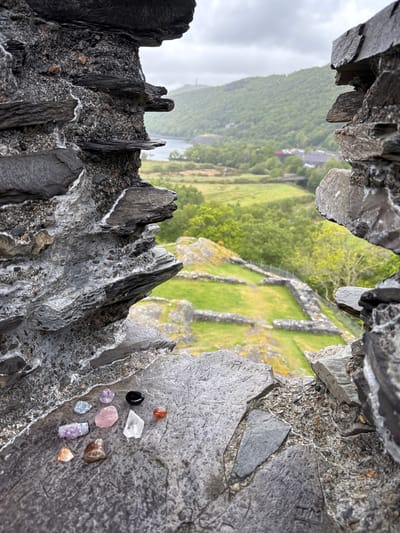 The image shows a view of a rural landscape through a gap in a rocky terrain. The foreground consists of jagged, gray rocks, some with visible crystalline or mineral deposits. A collection of small, colorful gemstones or crystals are resting on one of the rock surfaces. The landscape seen through the gap appears to be a green valley with fields, scattered buildings, and stone walls. The weather seems overcast, with a slightly hazy or misty quality to the scene. The city of Llanberis, United Kingdom is not evident in the photograph.
