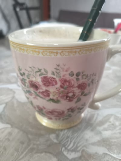 A close-up, slightly blurred shot shows a pink ceramic teacup adorned with a floral pattern of roses and green leaves. The teacup has a white rim with a yellow decorative band and rests on a saucer or tablecloth with a grey and white patterned texture. A dark green spoon with copper-colored accents is partially submerged in the foamy liquid within the teacup. The background is out of focus, with hints of furniture and possibly another object with a pink component. The lighting suggests an indoor setting, likely a home or cafe. There is no information to suggest this image is from Lisakovsk, Kazakhstan.