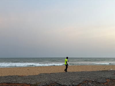 A lone figure in a neon shirt walks along a beach. The sky is overcast, and the sea stretches to the horizon, with small waves breaking on the sandy shore. A dark strip of pebbles or stones lies in the foreground. The man seems to be looking at a phone in his hand. The overall scene conveys a sense of solitude.