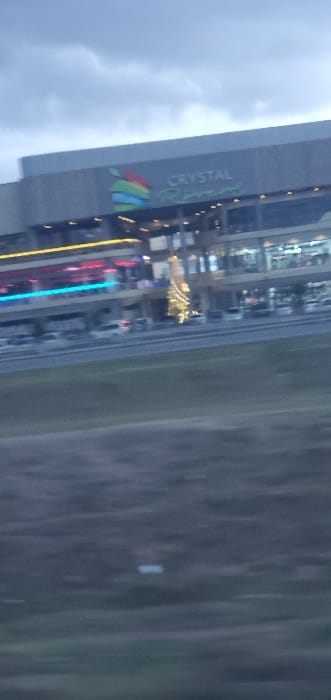 The image displays a multi-story commercial building, identified by the prominent "CRYSTAL Rivers" signage featuring a colorful, layered logo, situated in Kinanie ward, Kenya. The scene is captured at twilight or dusk under an overcast sky. The building's facade is illuminated with various lights, including horizontal red and blue strip lights on a lower floor, and yellow accent lighting. A large, conical, golden illuminated structure, resembling a Christmas tree, stands at ground level in front of the building. Below the building, a paved area, likely a parking lot, contains several indistinct vehicles, predominantly white. A dark-colored road separates this paved area from a grassy foreground. The entire image exhibits slight motion blur. No discernible individuals or explicit interactions are visible.