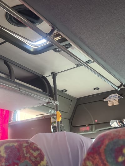 Interior shot of a bus. A metal handrail runs diagonally upward from the left, next to a window with a black rim. A hinged panel is visible behind the rail. Above are light gray ceiling panels. In the upper right, the dashboard is gray. A person wearing a white shirt and floral print sleeves stands in the foreground. The seat has a red covering.