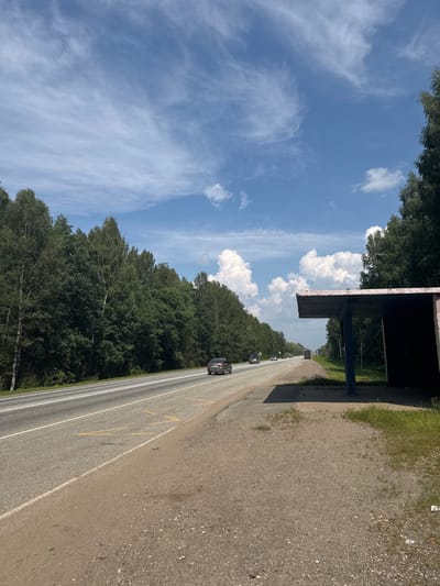 A paved road runs through a forest, with a bus stop on the right side of the road. The road has two lanes in each direction and is marked with yellow lines. Several cars are driving on the road. The bus stop is a simple structure with a concrete roof and two pillars. The forest is dense and filled with green trees. The sky is blue with white clouds. The location is Kabanov Mys, Russia.