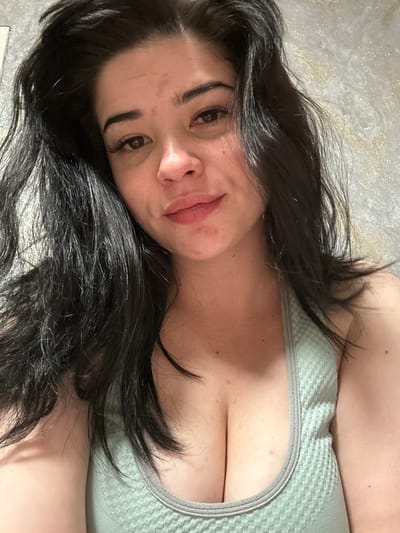 A close-up, waist-up selfie shows a young woman with long, dark hair framing her face. She is looking towards the camera with a slight smile. Her skin has a light complexion and some redness on her forehead. She is wearing a light green tank top with a textured pattern. The background appears to be a wall with a subtle, shimmering pattern. The image does not provide any information about the location in the city of Gavrilov Posad, Russia.