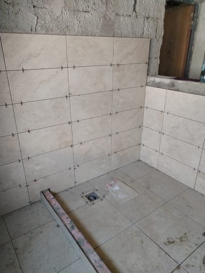The image depicts an interior scene, likely a bathroom or shower area under construction. The walls are partially tiled with light-colored, square tiles. Thin black lines, likely grout lines or guides for tiling, are visible between the tiles, with small plastic spacers inserted at the intersections. The rough concrete ceiling is visible above the tiled area. A rough concrete wall with exposed rebar is present on the left side of the image. On the right, there is a rough opening in the wall revealing a wooden surface, possibly a door or frame, and a window. The floor is also tiled with similar light-colored square tiles, some of which appear to be laid out. In the foreground, a metal leveling tool with a pink surface lies on the floor. A drain with a metal grate is visible on the floor, surrounded by tiles and spacers. A clear plastic bag is placed on the floor near the drain. The overall environment suggests an ongoing construction or renovation project. The location is identified as Lipănescu, Romania. There are no people visible in the image.
