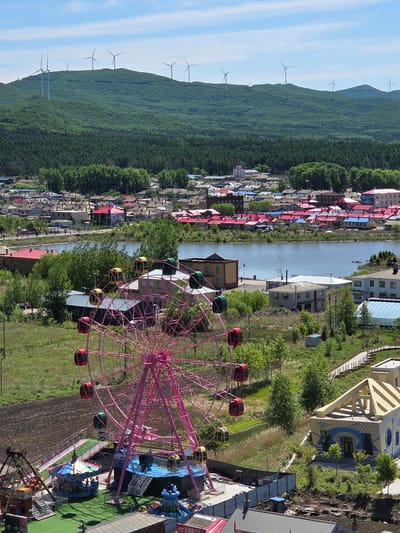 An aerial view captures a vibrant landscape in Jiejinkou, China, combining natural beauty with human development. In the foreground, a prominent pink Ferris wheel stands, adorned with multi-colored gondolas in red, yellow, and green, suggesting an active amusement park. Adjacent to the Ferris wheel are other amusement rides, one notably blue-themed, all situated on bright green artificial turf. To the right of the rides, a distinctive building with yellow and blue patterns features an arched design, possibly serving as an entrance or themed structure.

A calm river or canal gracefully flows horizontally across the midground, dividing the scene. On the left bank of the river, a sprawling village extends, characterized by numerous houses, many of which boast brightly colored roofs in shades of red and blue, adding a lively touch to the residential area. The right bank of the river is lined with more buildings and abundant green trees, blending into the park's perimeter.

The background is dominated by a majestic, densely forested mountain range under a clear blue sky with faint wisps of clouds. Along the mountain's ridgeline, several wind turbines are visibly spaced, indicating a wind farm utilizing the elevation. The scene is bathed in bright daylight, suggesting a clear, sunny day, enhancing the vibrant colors of the architecture and natural environment. People are implied to be present enjoying the amusement park attractions, although individual figures are small due to the high vantage point. Visible text, likely Chinese characters, appears on a red and white sign on a building near the amusement park, but it is illegible due to the distance and resolution.