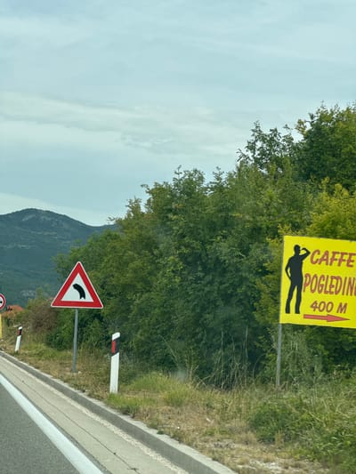 The image shows a roadside scene in Vidrovan, Montenegro. A winding road is visible in the foreground, with a concrete curb and grassy verge. Two road signs are present: a triangular warning sign with a black curve indicating a sharp bend to the left, and a yellow sign with a silhouette of a person drinking and text that reads "CAFFE POGLEDIINE 400 M," with a red arrow pointing to the right. In the background, lush green trees and hills create a natural landscape. The sky is overcast with clouds.