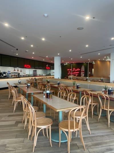 The image depicts the interior of a restaurant in Surfers Paradise, Australia. The restaurant has a modern and clean aesthetic with light wooden floors and tables. Red neon signs on the wall display the words "PIZZA" and "PASTA," indicating the restaurant's specialization. The seating arrangement includes several wooden chairs and banquette seating along the walls. The tables are set with condiments, including bottles of oil and vinegar. In the background, a counter area is visible with kitchen equipment and what appears to be a food preparation space. Another neon sign in the background reads "I'm in Pasta Paradise." The overall scene suggests a casual dining environment.