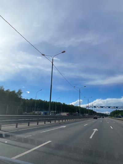 The image depicts a highway in Новосёлки, Russia on a sunny day with a blue sky and scattered clouds. The highway has multiple lanes marked with white lines and arrows indicating direction. Several vehicles are visible in the distance. Along the side of the highway, there is a metal guardrail and trees. Overhead, there are streetlights lining the road.