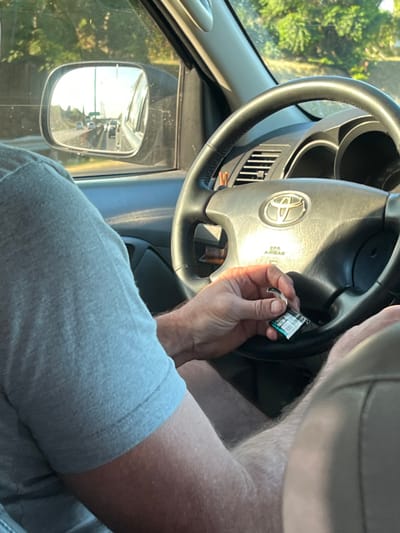 Inside a car, an individual holds a small package of gum near the Toyota steering wheel. The steering wheel shows the Toyota logo. The side mirror reflects the road and adjacent trees outside. The person is wearing gray clothing. The interior of the car is gray and black.
