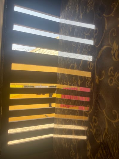 The image displays an indoor perspective looking through a multi-slatted window or shutter in Dzhidu, Nigeria. The left side features dark horizontal slats interspersed with bright light. Through the openings, an exterior environment is visible, characterized by a light blue sky, a greenish corrugated metal roof, and razor wire positioned along a boundary. Below, laundry, including yellow, white, and pink garments, hangs on an outdoor clothesline. The right side of the image shows an interior wall adorned with dark brown wallpaper featuring golden floral and scroll patterns. Sunlight projects distinct, bright horizontal bands onto this wallpaper, with some bands exhibiting color reflections from the exterior laundry. No individuals are present in the scene.