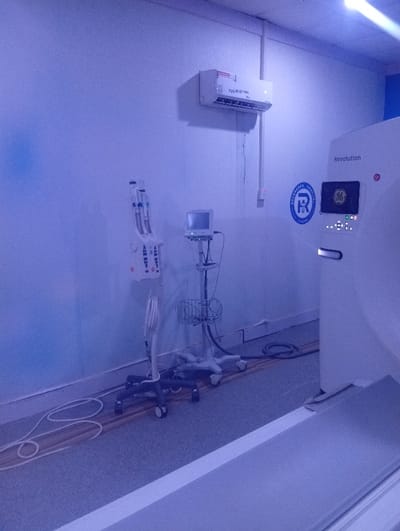 The image depicts the interior of a medical diagnostic room within Heritage Hospital, located in Okengwe - Obehira, Nigeria. The scene is illuminated by artificial light, casting a distinct blue hue across the environment.
On the right side of the frame, a large, white General Electric (GE) "Revolution" computed tomography (CT) scanner is prominent. The machine features an active display screen showing green and yellow graphical elements and bears the blue circular logo of "Heritage Hospital."
To the left of the CT scanner, two additional medical devices on wheeled stands are present. The leftmost stand supports a dual-head contrast injector with two clear cylindrical chambers at its top. Adjacent to it, another wheeled stand holds a small medical monitor and a metal wire basket positioned beneath the screen.
The walls are light-colored and unadorned. A white LG split-unit air conditioner is mounted high on the wall above the ancillary equipment, with electrical conduits descending from it. The floor is light gray, with multiple white and black electrical cables visible, some routed along a brown trim strip at the base of the wall. No individuals are present in the room, and no discernible actions or interactions are occurring.
