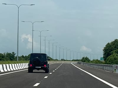 A black Mercedes-Benz G-Wagon is driving on an asphalt road in სუფსის თემი, Georgia. The road has white lane markings and is bordered by a black and white striped barrier on the left and a concrete barrier with red and white reflectors on the right. A long line of streetlights stretches into the distance along the left side of the road. Lush green trees and vegetation are visible on the sides of the road. The sky is overcast with some clouds. The G-Wagon has a spare tire mounted on the rear door and a license plate that is partially visible. The scene appears to be a highway or a major road.