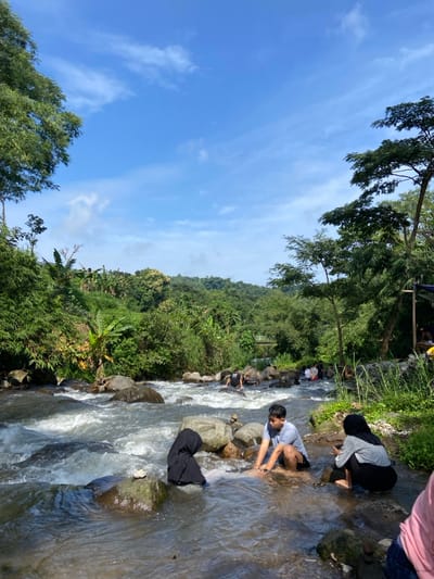 A stream flows diagonally through the frame, flanked by lush green vegetation on its left bank and rocky terrain on the right. Several people are near or in the water. One person in a hijab and another in a light-colored shirt are near the stream. Others can be seen further upstream. Trees line the upper edge of the greenery. The sky above is mostly clear, with scattered clouds.