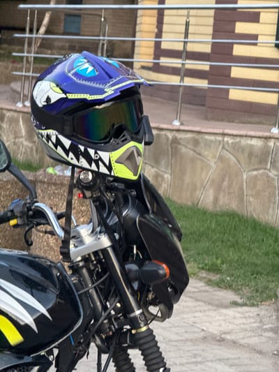 The image depicts a motorcycle with a helmet placed on its handlebars. The helmet is blue with a graphic design resembling shark teeth and fins. It has a reflective visor with a rainbow sheen. The motorcycle is black and has silver accents on its fuel tank. In the background, there is a building with horizontal striped siding and a metal railing. Green grass and a paved surface are also visible. The scene appears to be outdoors, possibly in a residential area or a parking lot. The location is identified as Piatykhatky, Ukraine.