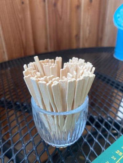 The image shows a close-up of a glass filled with wooden skewers. The skewers are arranged neatly and appear to be unused. The glass is clear with a ribbed texture around its base. The background features a wooden surface to the left, possibly a table or wall, and a dark, metallic mesh surface to the right, which could be part of outdoor furniture. A small portion of a green object with yellow markings, possibly a ruler or measuring tape, is visible in the upper right corner. The markings on the green object include the numbers "25" and "24". The lighting suggests it might be daytime, and the setting appears to be outdoors or in a well-lit indoor space. There is no indication of people or any specific activity taking place. The focus is entirely on the skewers and the glass container.